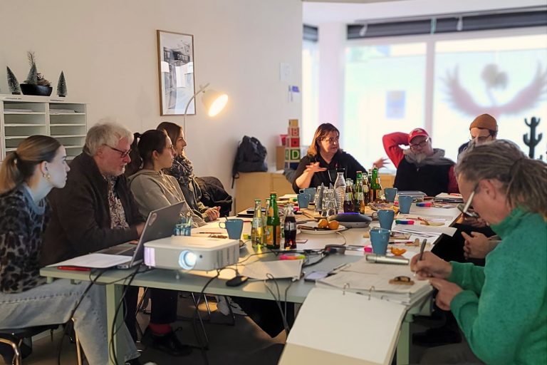 Treffen der LAG Spielmobile NRW. Acht erwachsene Menschen sitzen um einen großen Tisch und unterhalten sich. Auf dem Tisch sind Dokumente und Getränke verteilt.